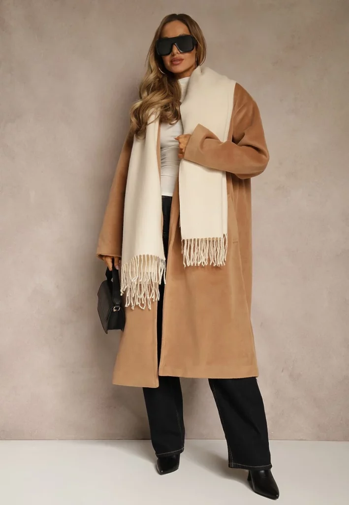 Modelka prezentuje beżowy długi płaszcz z szalikiem i okularami

