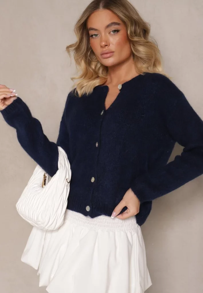 Modelka prezentuje zestaw z granatowym swetrem typu kardigan, białą spódniczką balloon i białą pikowaną torebką do ręki.