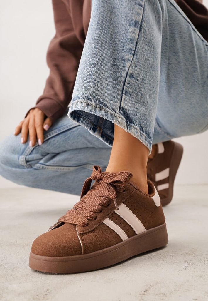 Modelka prezentuje brązowe sznurowane sneakersy spotowe na grubej podeszwie z białymi paskami. 