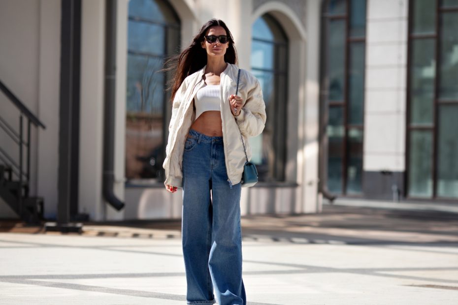 Modelka prezentuje białą bomberkę w połączeniu z krótkim topem i szerokimi jeansami.
