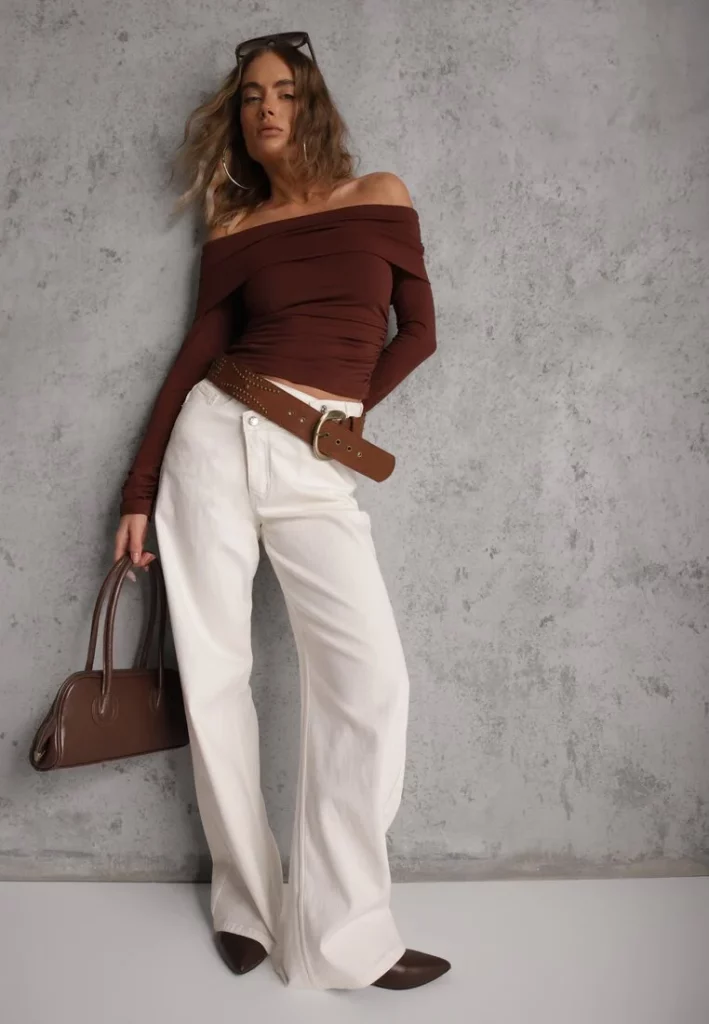 Modelka prezentuje brązową bluzkę z marszczeniami z odkrytymi ramionami. Do tego ma na sobie białe szerokie spodnie i duży pasek. 