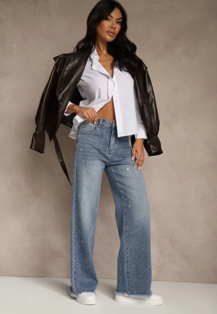 Modelka prezentuje zestaw z szerokimi jeansami w odcieniu niebieskiego, które są ozdobione cyrkoniami. Do tego koszula i czarna skórzana kurtka. 