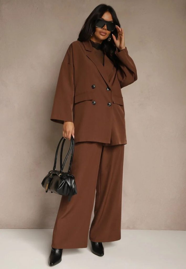 Modelka prezentuje brązowy total look z garniturem oversize, torebką bowling, botkami na szpilce i okularami przeciwsłonecznymi.