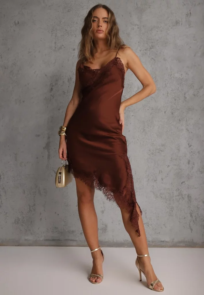 Modelka prezentująca brązową sukienkę asymetryczną na ramiączkach ze złotymi dodatkami - buty, torebka, biżuteria. 
