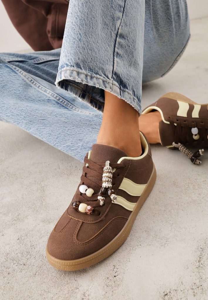 Modelka prezentuje brązowe sneakersy z białymi paskami i koralikami w towarzystwie jasnych jeansów 