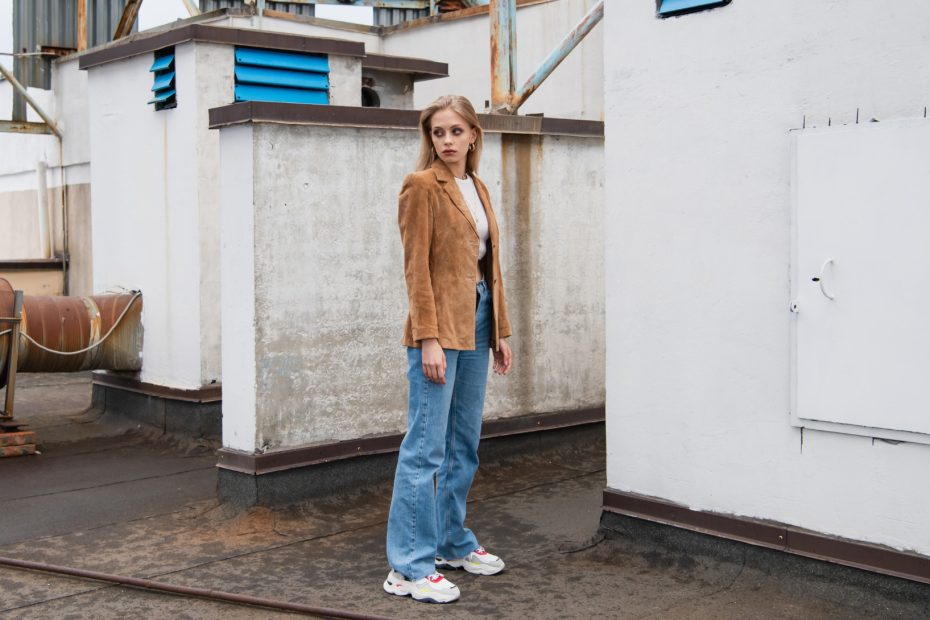 Modelka pozuje w jeansach i zamszowej marynarce na pofabrycznej, industrialnej scenerii.