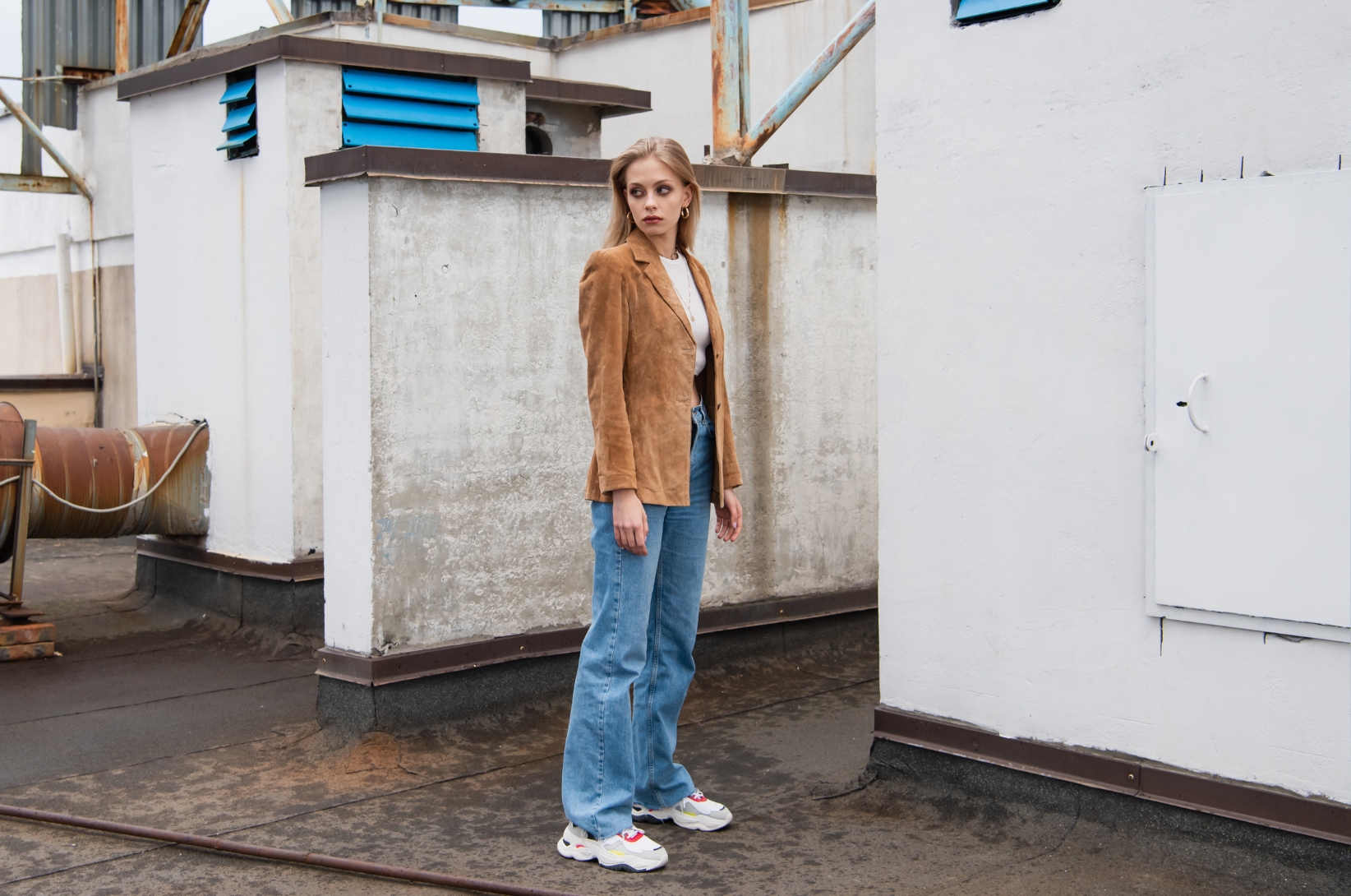 Modelka pozuje w jeansach i zamszowej marynarce na pofabrycznej, industrialnej scenerii.