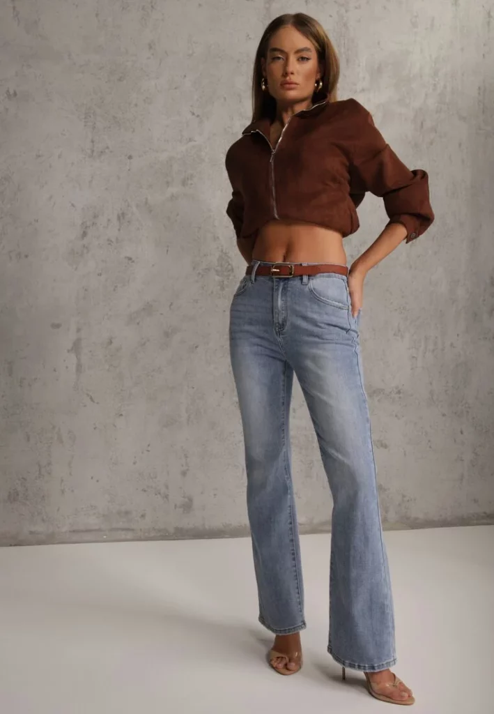 Modelka prezentuje jeansy dzwony o fasonie bootcut z wyoskim stanem. Nosi dodatkowo pasek i kurtkę brązową. 