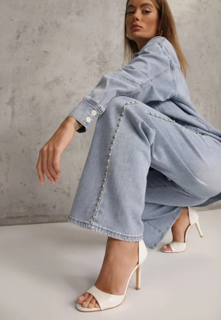 Modelka pozuje kucając i prezentuje szerokie spodnie z nitami w jasnym odcieniu błękitu.